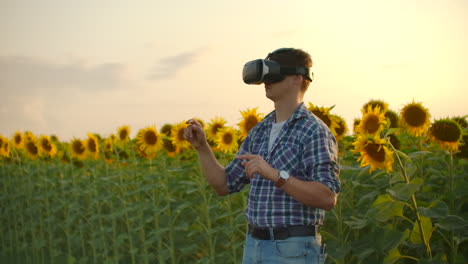 A-young-farmer-in-plaid-shirt-and-jeans-uses-VR-glasses-on-the-field-with-sunflowers-for-3D-moderling.-These-are-modern-technologies-in-summer-evening.